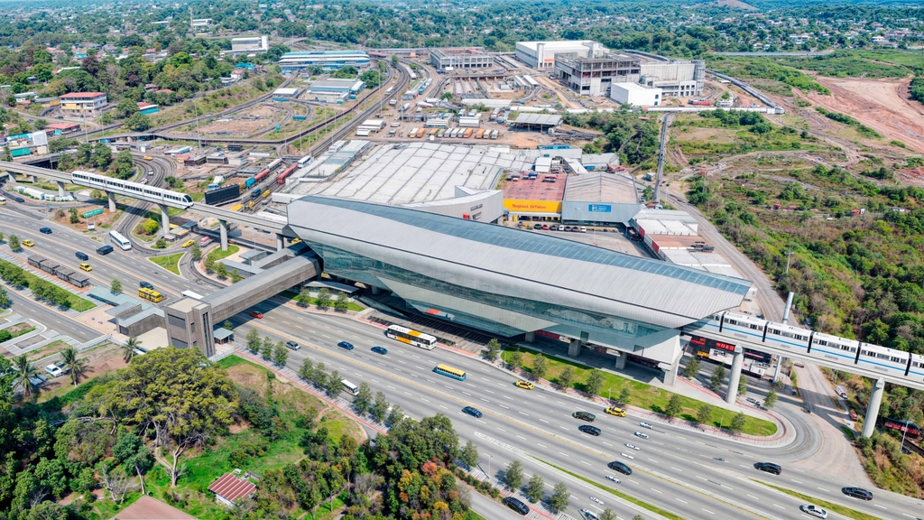 Vista aérea de una estación elevada de metro, con un tren sobre el viaducto y una autopista en primer plano.