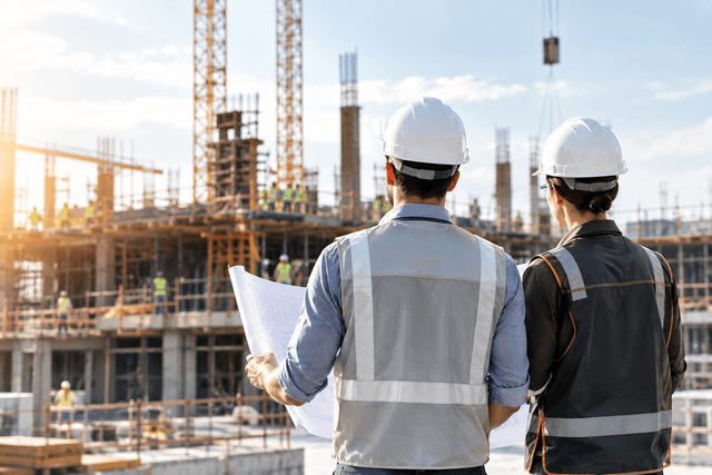 Dos técnicos con casco y chaleco revisan un plano frente a un edificio en construcción.